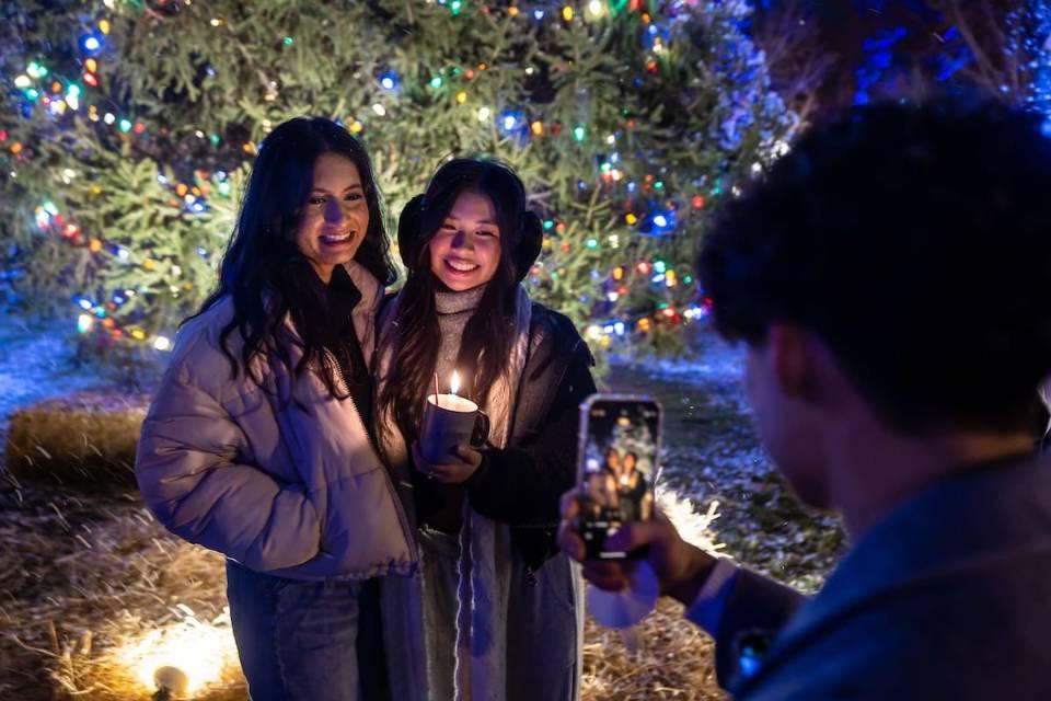 两名身着冬衣的博彩网址大全女学生在学校的圣诞树前摆姿势拍照, 哪个被彩灯覆盖. 一个学生拿着一个杯子，里面有一支点燃的蜡烛. 另一名学生站在前景，用智能手机拍照.